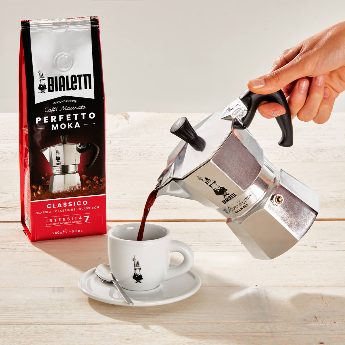 "Bialetti Moka pot pouring coffee into a cup with a Bialetti coffee bag in the background."