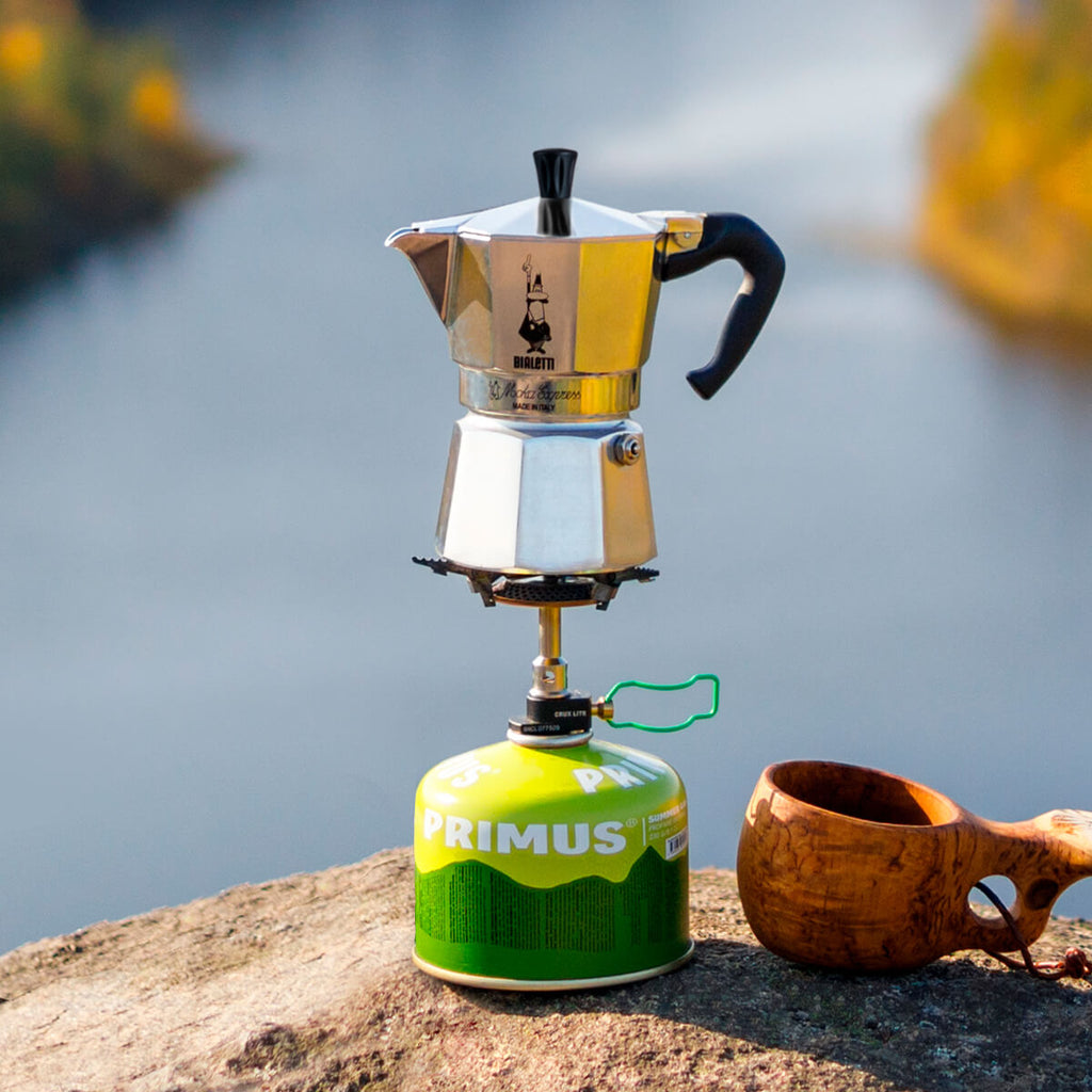 "Portable coffee maker on a Primus stove with a wooden cup by a body of water."