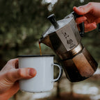 "Person pouring coffee from a moka pot into a white mug with a blurred natural background"