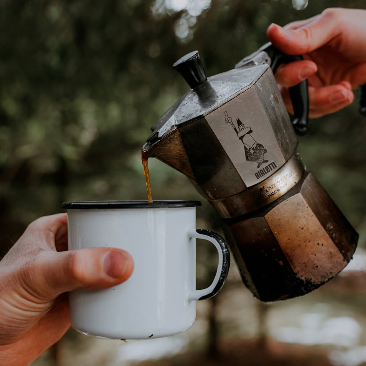 "Person pouring coffee from a moka pot into a white mug with a blurred natural background"